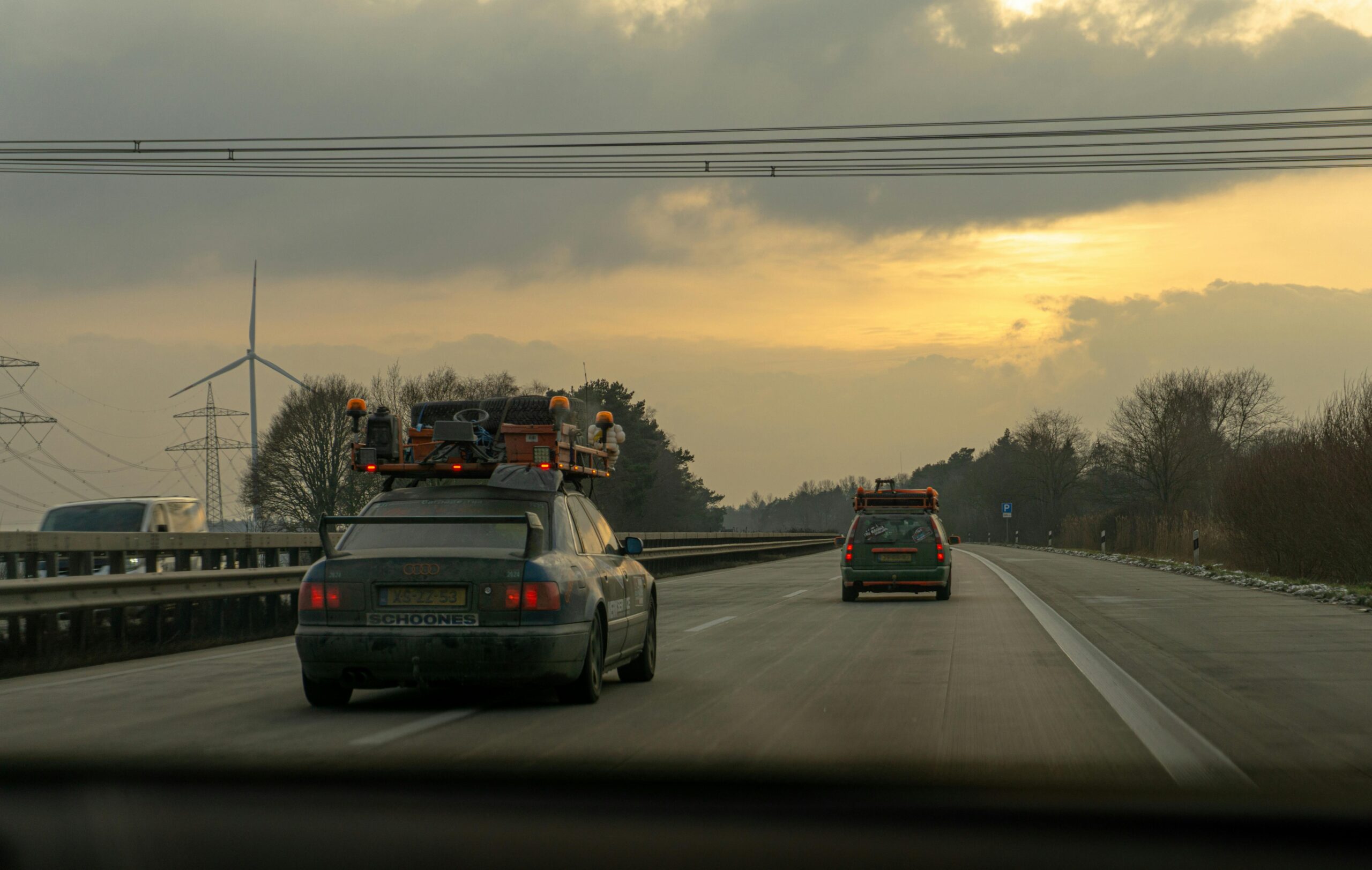 Zwei Autos auf einer deutschen Autobahn bei Sonnenuntergang mit Windrädern und Stromleitungen im Hintergrund.
