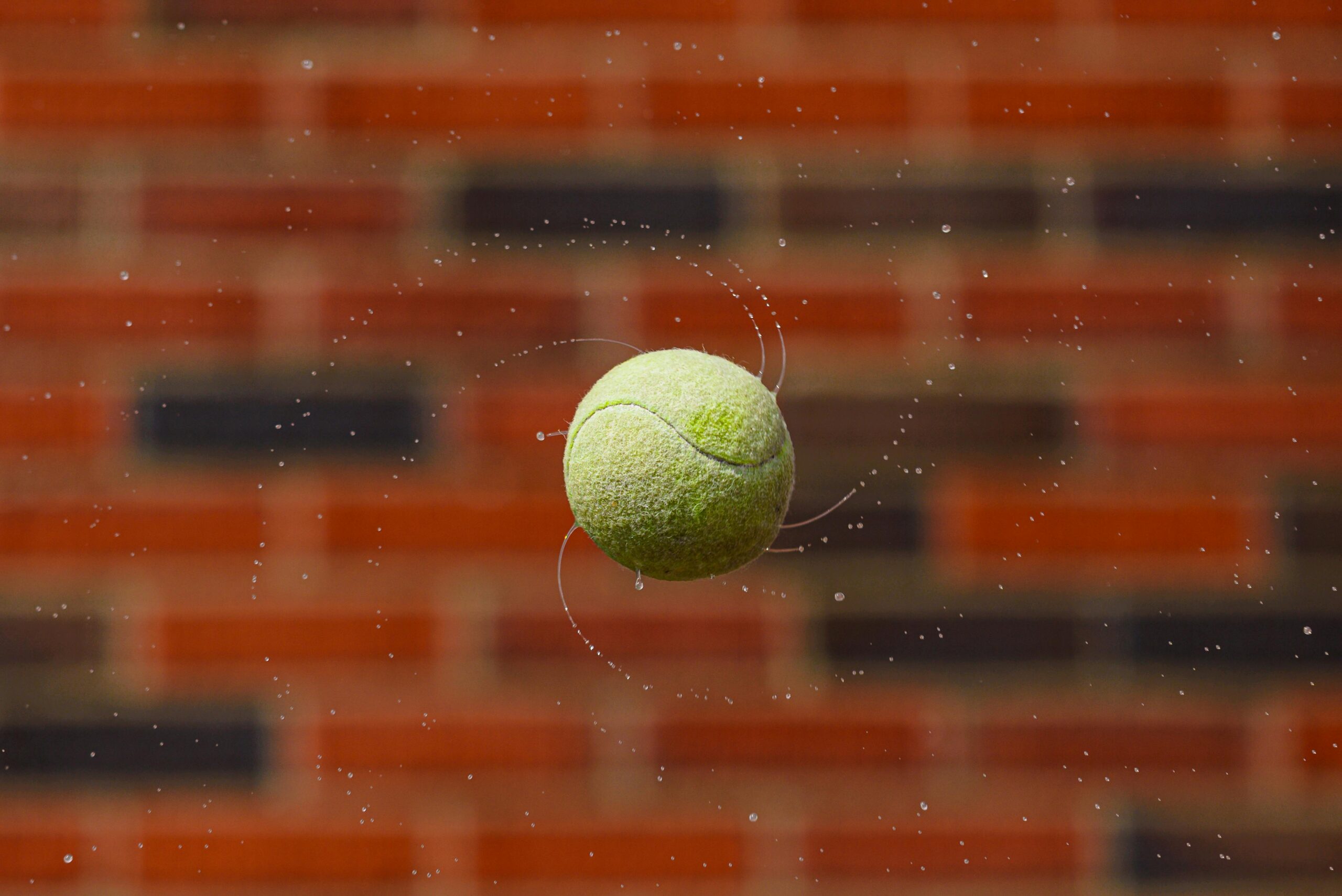Nahaufnahme eines nassen Tennisballs, der im Freien mitten im Spin vor einer Backsteinmauer aufgenommen wurde.